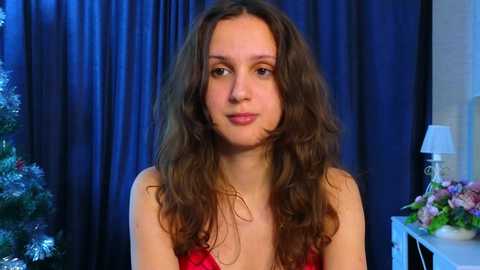 Media: Video of a young woman with long, wavy brown hair and fair skin, wearing a red sleeveless top. She stands against dark blue curtains, with a Christmas tree and floral arrangement in the background.