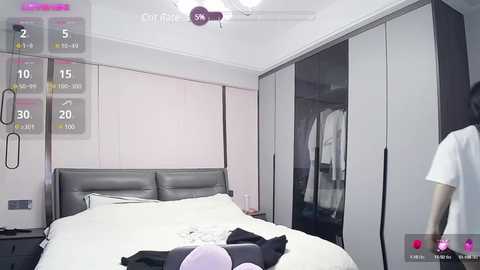 Media: Video of a minimalist bedroom with a large gray leather bed, white sheets, and a white wardrobe with a hanging shirt. The room is bright, modern, and clean.