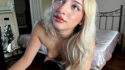 Media: Video of a young woman with long, platinum blonde hair, light skin, and full lips, wearing a black strapless top, leaning over a bed in a cozy, dimly-lit bedroom.