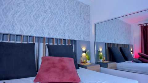 Media: A video of a modern bedroom with a large, plush red headboard, gray patterned wallpaper, a mirrored wall, and bedside lamps with green plants.