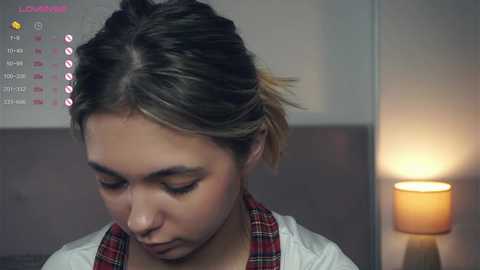 Media: Video of a young woman with light brown skin and short, dark hair, wearing a red and white plaid shirt, sitting indoors, with a digital clock displaying time and social media icons in the background.