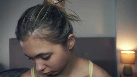 Media: Video of a young woman with fair skin, light brown hair in a messy bun, wearing a yellow tank top, sitting on a bed, focusing intently on a book. Background features a beige wall and a lit lamp.