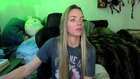 Media: Video of a young, slim, blonde woman with long hair, wearing a grey t-shirt, seated on a black chair in a cluttered room with a green-lit background.