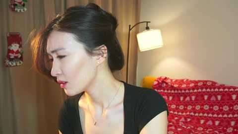 Video of an East Asian woman with fair skin and dark hair in a side ponytail, wearing a black top, standing indoors. She is illuminated by a soft lamp with a white shade.