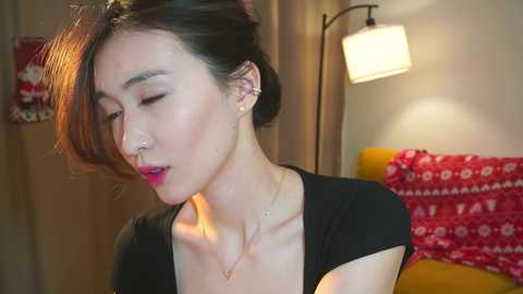 Video of an Asian woman with medium-length, dark brown hair styled in a side part, wearing a black top and gold necklace. She appears serene, with closed eyes and subtle makeup. Background features a yellow couch and festive red blanket.