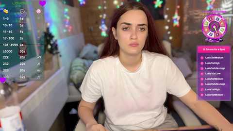 Media: Video of a young woman with long brown hair, wearing a white t-shirt, sitting in a dimly lit room with colorful Christmas lights.