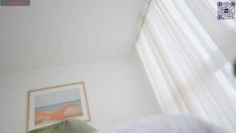 Media: A video showing a minimalist bedroom with white walls, sheer curtains, and a framed abstract painting featuring pastel colors. The bed, covered in white sheets, is partially visible on the left.