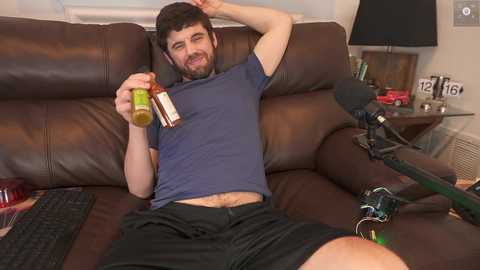 Media: Video of a man with a beard, blue shirt, and black shorts, lounging on a brown leather couch, drinking a green drink, holding a book, and using a microphone for a YouTube livestream.