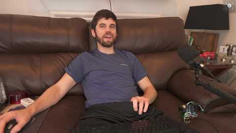 Media: Video of a bearded man with short dark hair, wearing a blue t-shirt and black shorts, reclining on a brown leather couch. He's holding a game controller. A black microphone and a red lamp are visible in the background.