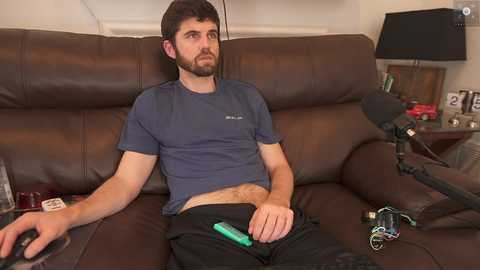 Media: Video of a bearded, fair-skinned man with short dark hair, wearing a blue t-shirt, black pants, and holding a green controller, sitting on a brown leather couch in a dimly-lit living room.