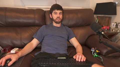 Media: Video of a bearded, dark-haired man in a gray T-shirt, sitting on a brown leather couch, wearing headphones, with a gaming controller in his lap.