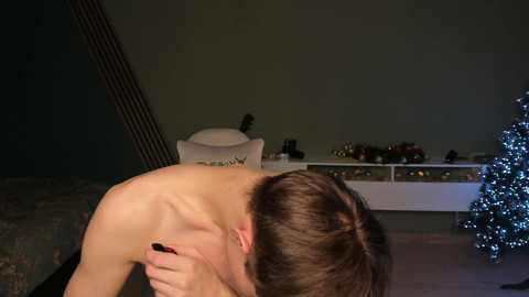 Media: Video of a shirtless, fair-skinned man with short brown hair, covering his face with his right hand, standing in a dimly lit room with a white chair and Christmas decorations.