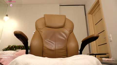 Media: Video of a beige leather recliner chair in a minimalist, beige-walled bedroom with a white door and a bed with a pink blanket in the background.