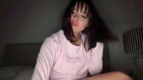 Media: Video of an Asian woman with long black hair, wearing a light pink hoodie, smiling, sitting on a bed with a gray headboard and a black and white lamp.