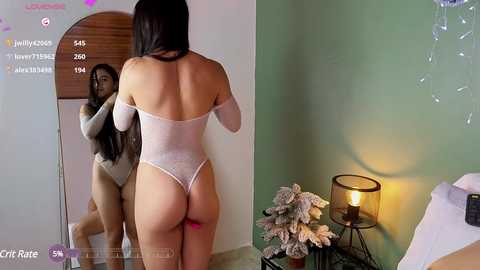 Media: Video of a woman in a white, backless, sheer bodysuit, adjusting her hair in a mirror, green walls, potted plants, and a lit lamp.