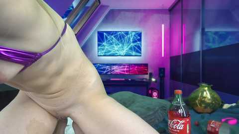 Media: Video of a light-skinned, slender woman in a metallic purple bikini, sitting on a bed in a modern room with purple and blue lighting, a green glass bottle, and a Coca-Cola can.