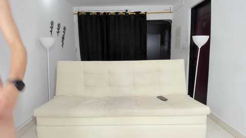 Media: Video of a minimalist, modern living room with a white leather couch, black curtains, white wall-mounted lamp, and a television remote on the couch.