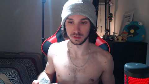 Media: Video of a shirtless young man with a grey beanie, sitting in a red and black gaming chair. Background includes a blue plush toy and a patterned pillow.