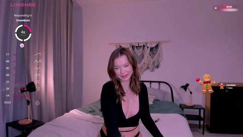 Media: A video of a smiling woman with long, wavy brown hair, wearing a black top, standing in a dimly lit bedroom with a bed, a wall hanging, and a nightstand.