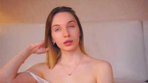 Media: Video of a fair-skinned, slender woman with shoulder-length, light brown hair, wearing a white off-the-shoulder top, posing against a beige textured wall, looking thoughtful, with a delicate necklace.