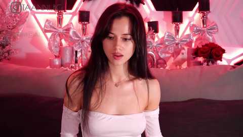 Media: Video of a young woman with long black hair, wearing a white strapless top, standing in a dimly lit room with red and silver decor, including floral arrangements and candles.