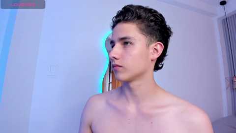 Media: Video of a young, shirtless, light-skinned man with curly dark hair, standing against a white wall with a blue light strip.