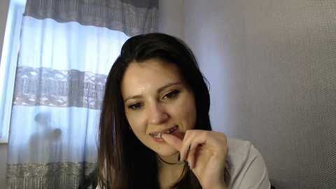 Media: Video of a young woman with straight, dark brown hair, fair skin, and a light beige top, smiling and gently biting her finger, set against a background of a sheer white curtain with lace trim.