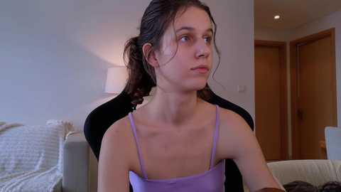 Media: Video of a young woman with light skin and dark hair in a ponytail, wearing a lavender spaghetti-strap top, sitting on a black gaming chair, in a modern, softly lit living room.