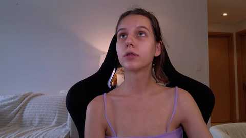 Media: Video of a young woman with light skin, brown hair, and a slim build, wearing a lavender spaghetti-strap top, sitting on a black gaming chair in a sparsely decorated bedroom with a white bed and wooden door.