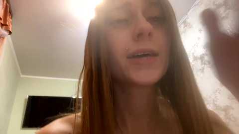 Media: Video of a young woman with long, straight brown hair, fair skin, and a nose piercing, taken from a low angle, showing her upper body and face. She appears to be indoors, with a wall and a TV visible in the background.