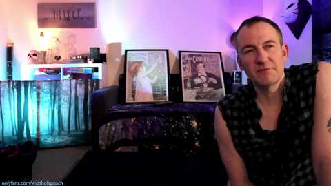 Media: Video of a man in a black shirt, sitting in a dimly lit room with a purple and blue lighting ambiance. Behind him, two framed magazine covers hang on a wall, featuring a woman in a white dress and a man in a suit.