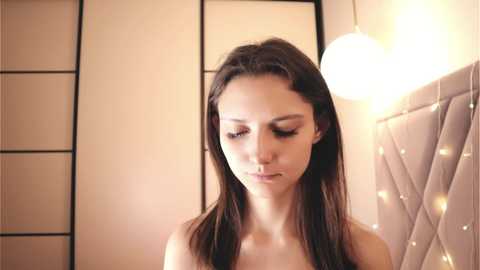 Video of a young woman with long, straight brown hair, wearing minimal makeup, standing indoors near a white, softly lit, round lamp and a beige quilted headboard.