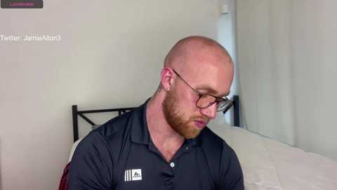 Video of a bald, bearded man with glasses, wearing a black polo shirt, sitting on a white bed in a minimalist room.