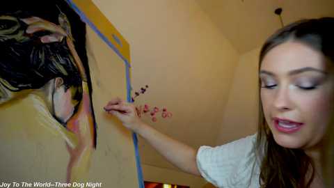 Media: Video of a woman with long brown hair painting a mural of a woman in a yellow room, adding small red dots.