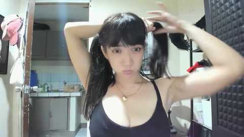 Media: Video of an Asian woman with long black hair in pigtails, wearing a black tank top, standing in a cluttered bathroom with a white sink and mirror.