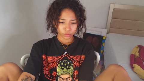 Media: Video of a young Black woman with curly hair, wearing a black t-shirt with a graphic design, sitting on a bed, looking contemplative.