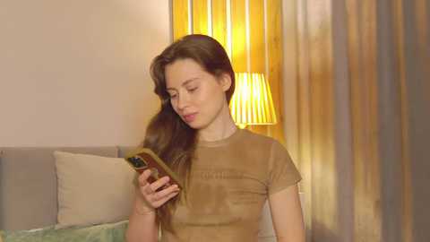 Media: Video of a young woman with long brown hair, wearing a beige t-shirt, holding a smartphone, in a warmly-lit bedroom with a beige bed and a yellow lampshade.