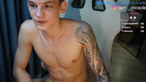 Media: Video of a shirtless young man with a muscular build, light skin, and intricate sleeve tattoos, seated indoors.