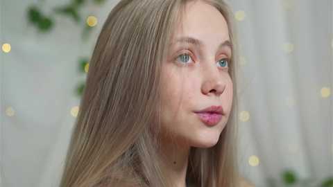 Media: Video of a young Caucasian woman with long, straight blonde hair, blue eyes, fair skin, and pink lips. She is wearing a brown top. Background is softly blurred with greenery and white curtains.