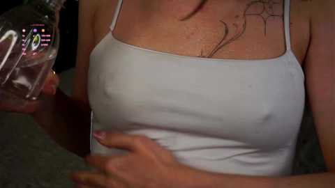 Media: A close-up video of a light-skinned woman with a tattoo on her neck, wearing a white tank top, drinking from a glass bottle, with a blurred background.