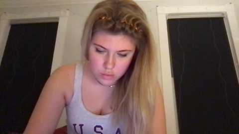 Media: Video of a young blonde woman with long hair styled in a high ponytail, wearing a grey \"USA\" tank top, looking down, in a dimly-lit room with closed blinds.