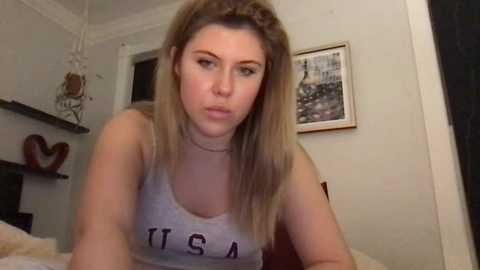 Media: A video of a young woman with long, straight, light brown hair, wearing a gray tank top with \"USA\" in purple letters. She puckers her lips in a playful manner, leaning forward. The background features a framed picture and a dark shelf with decorative items.