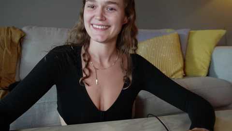 Media: Video of a smiling Caucasian woman with long brown hair, wearing a black, plunging V-neck top, sitting on a light gray couch adorned with yellow and mustard pillows.