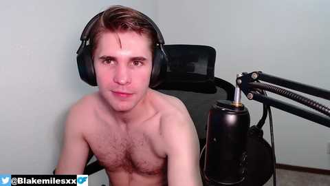 Media: Video of a shirtless, fair-skinned, young man with short brown hair wearing headphones, sitting at a desk microphone in a plain room.