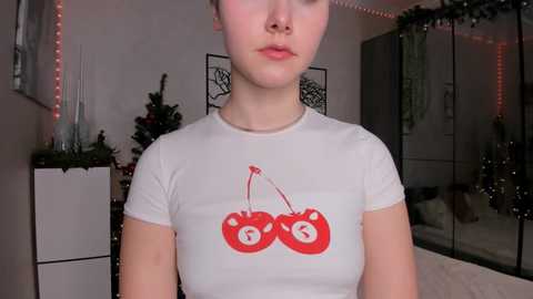 Media: Video of a young Caucasian woman with fair skin and light brown hair, wearing a white T-shirt with red cherry print and numbers, in a festive, dimly lit room with Christmas decorations.