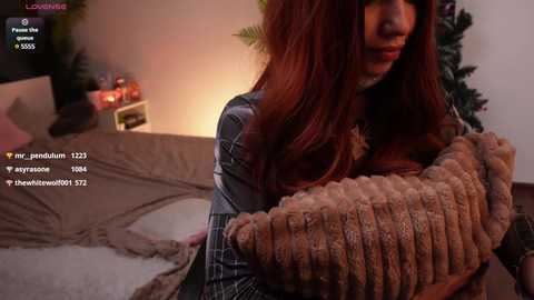 Media: Video of a woman with long, auburn hair, wearing a plaid shirt, cuddling a brown, furry blanket in a cozy bedroom with a Christmas tree.