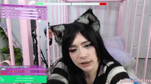 Media: Video of a pale-skinned woman with long black hair, wearing cat ears, a striped sweater, and a serious expression. Background shows a pink cage with green plants, a pillow, and a computer screen displaying a \"Crimson Cat\" game.