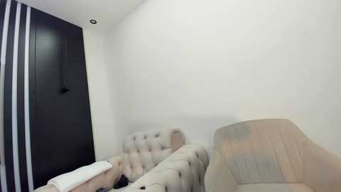 Video of a minimalist, dimly-lit bedroom with a beige tufted headboard, a beige armchair, a black-and-white striped curtain, and a dark wooden door on the left.