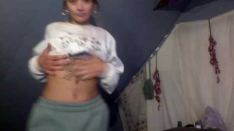 Media: A blurry video of a woman lifting her white crop top, revealing her tattooed midriff, wearing gray sweatpants. Background includes a dark wall, hanging red flowers, and a white curtain.