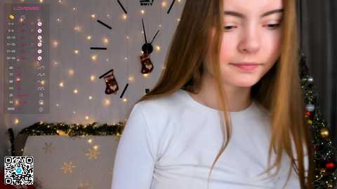 Media: Video of a young Caucasian woman with long, straight brown hair, wearing a white long-sleeved top, standing indoors near a decorated Christmas tree with golden lights and baubles.